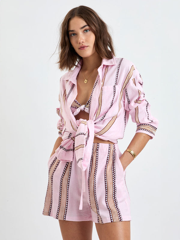 Woman Standing wearing lemlem MARIAM Shirt featuring detailed bands of deep burgundy, bronze, and pink stripes beautifully placed on a light cream 