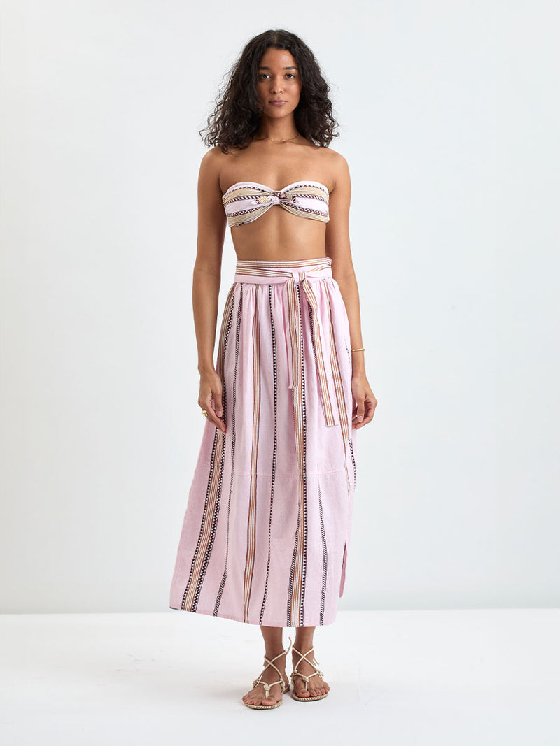 Woman Standing wearing lemlem AVA Bandeau Top and TOLA Skirt featuring detailed bands of deep burgundy, bronze, and pink stripes beautifully placed on a light cream 