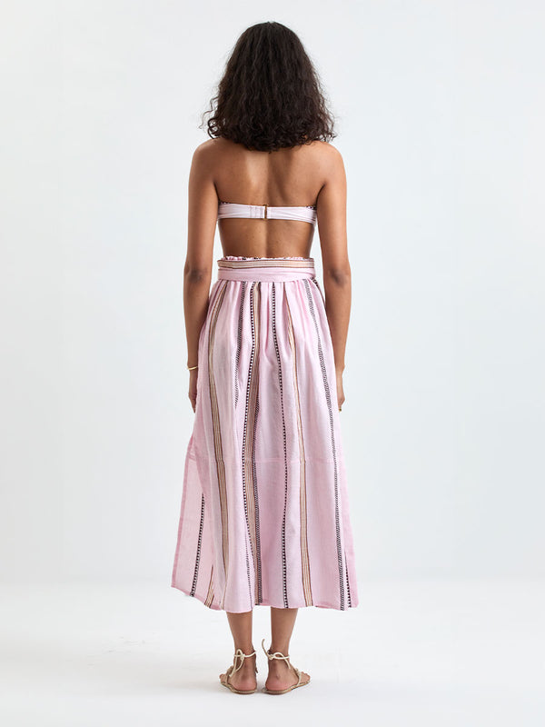 Back of a Woman Standing wearing lemlem AVA Bandeau Top and TOLA Skirt featuring detailed bands of deep burgundy, bronze, and pink stripes beautifully placed on a light cream 