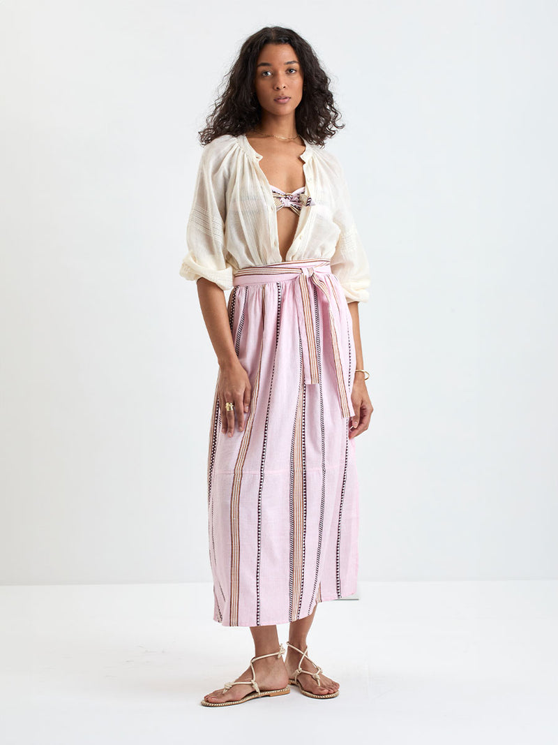 Woman Standing wearing lemlem MITA Blouse in cream color  and TOLA Skirt featuring detailed bands of deep burgundy, bronze, and pink stripes beautifully placed on a light cream 