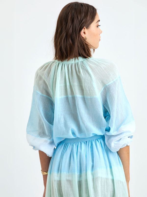 Back of a Woman standing wearing lemlem MITA Blouse featuring  light, gauzy fabric with a beautiful color block of light blue, aqua, mint, soft green and cream