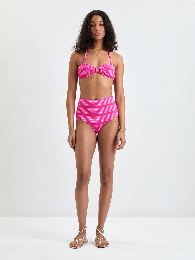 Woman Standing Wearing lemlem  AVA Bandeau Top and ELSI High Waist Bottom featuring vibrant bright pink stripes accented with ivory dots, all set against a bold fuchsia background