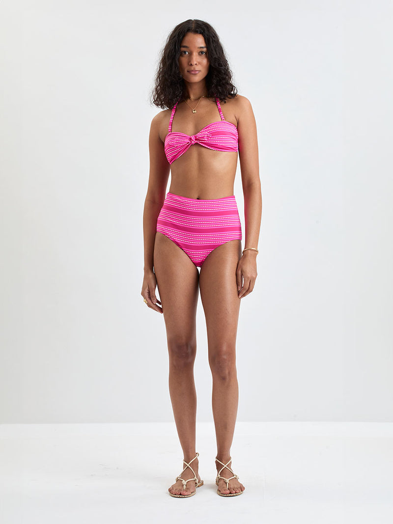 Woman Standing Wearing lemlem  AVA Bandeau Top and ELSI High Waist Bottom featuring vibrant bright pink stripes accented with ivory dots, all set against a bold fuchsia background