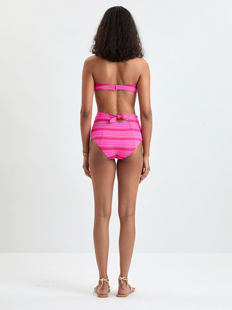 Back of a Woman Standing Wearing lemlem  AVA Bandeau Top and ELSI High Waist Bottom featuring vibrant bright pink stripes accented with ivory dots, all set against a bold fuchsia background