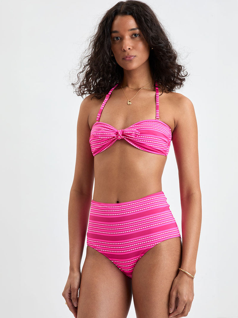 Woman Standing Wearing lemlem  AVA Bandeau Top and ELSI High Waist Bottom featuring vibrant bright pink stripes accented with ivory dots, all set against a bold fuchsia background