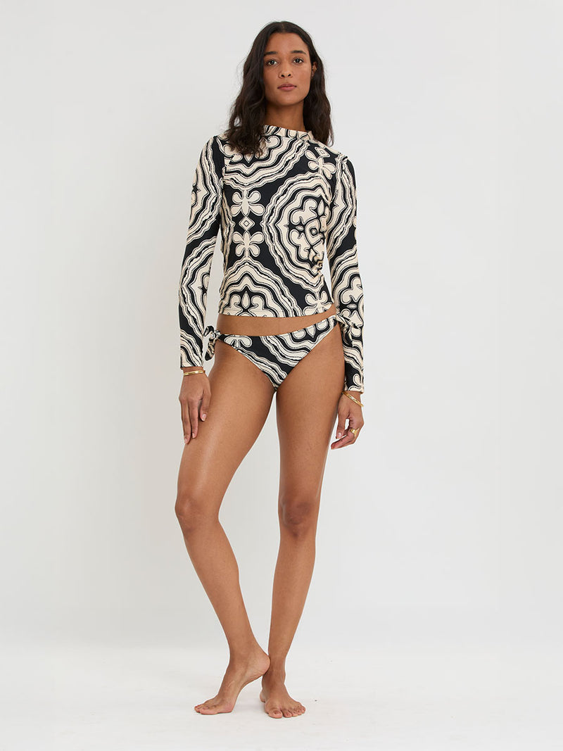 Woman standing wearing lemlem AZEB Rash Guard and LUCY Side Tie Bottom featuring ancient inspired pattern in creamy ivory perfectly grounded by rich black for a striking and timeless look.