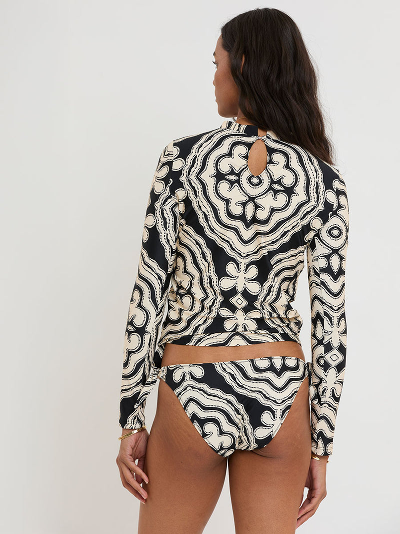 Back of a Woman standing wearing lemlem AZEB Rash Guard and LUCY Side Tie Bottom featuring ancient inspired pattern in creamy ivory perfectly grounded by rich black for a striking and timeless look.