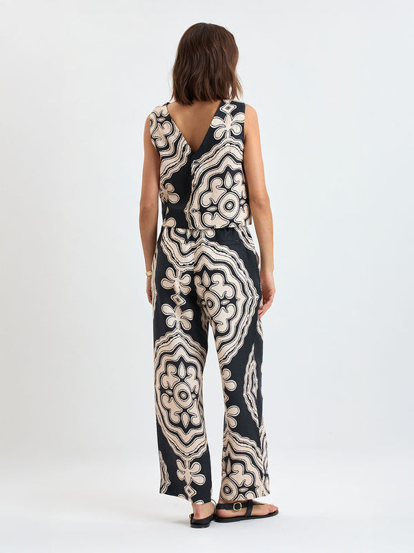 Back of a woman standing wearing lemlem DIRARRA Pants featuring ancient inspired pattern in cream color on black background and a matching tank top