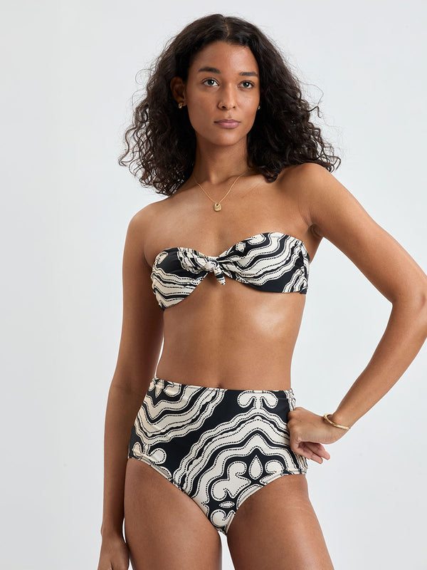 Woman standing wearing lemlem AVA Bandeau Top and ELSI High Waist Bottom featuring ancient inspired pattern in creamy ivory perfectly grounded by rich black for a striking and timeless look.