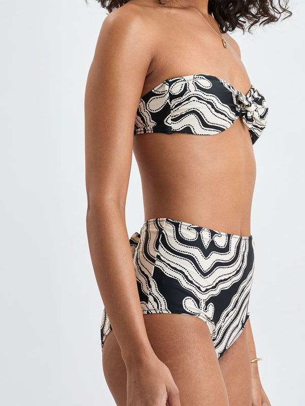 Side shot of a Woman standing wearing lemlem AVA Bandeau Top and ELSI High Waist Bottom featuring ancient inspired pattern in creamy ivory perfectly grounded by rich black for a striking and timeless look.