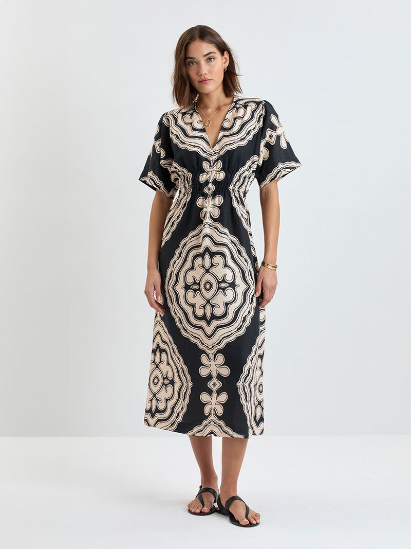 Woman Standing wearing lemlem linen HERMONA Plunge Dress  featuring  a custom-designed print inspired by ancient architectural patterns in cream and black colors