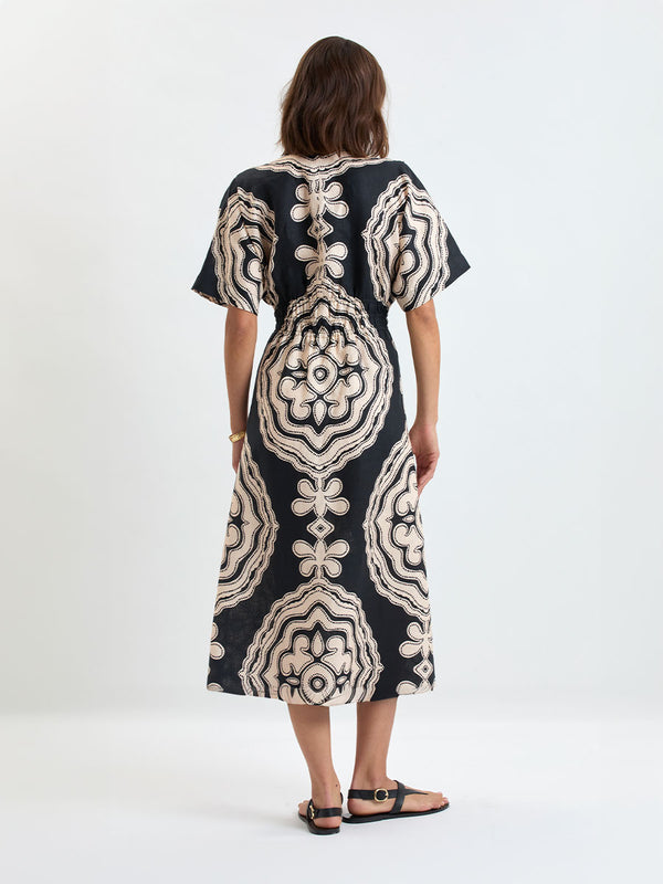 Woman's back shot Standing wearing lemlem linen HERMONA Plunge Dress  featuring  a custom-designed print inspired by ancient architectural patterns in cream and black colors