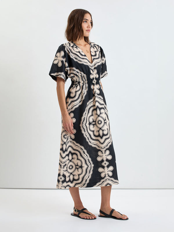 Side shot of a Woman Standing wearing lemlem linen HERMONA Plunge Dress  featuring  a custom-designed print inspired by ancient architectural patterns in cream and black colors