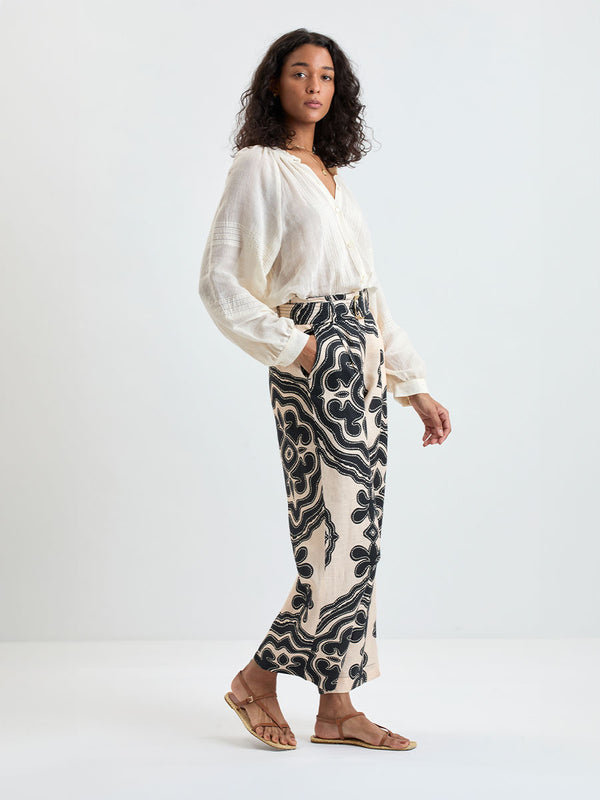 Woman Standing wearing lemlem MITA Blouse in cream color and DIARRA Pants featuring ancient inspired pattern in black color on cream background