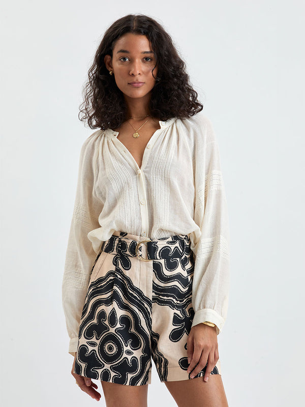 Woman Standing wearing lemlem MITA Blouse in cream color and FAIZAH Shorts featuring ancient inspired pattern in black color on cream background