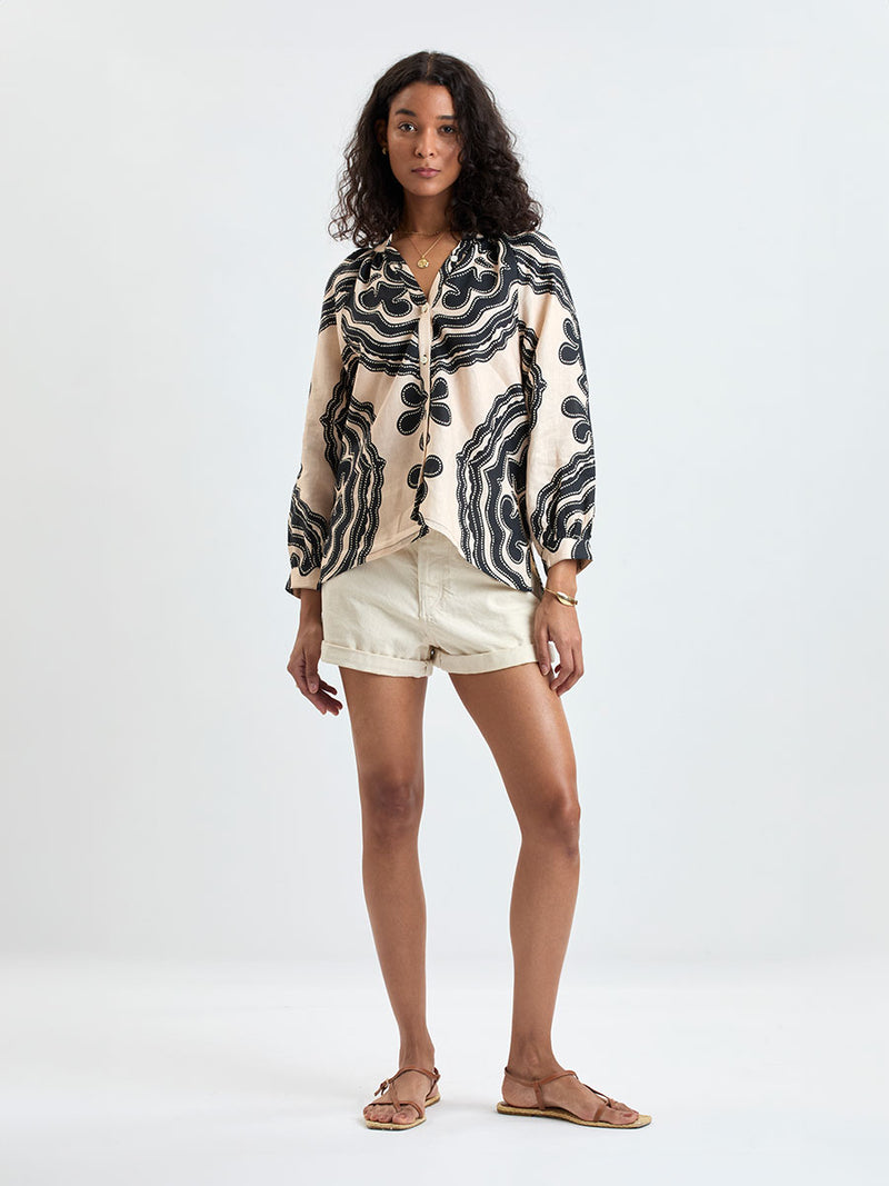 Woman Standing wearing lemlem MITA Blouse featuring ancient inspired pattern in black color on cream background and beige denim shorts