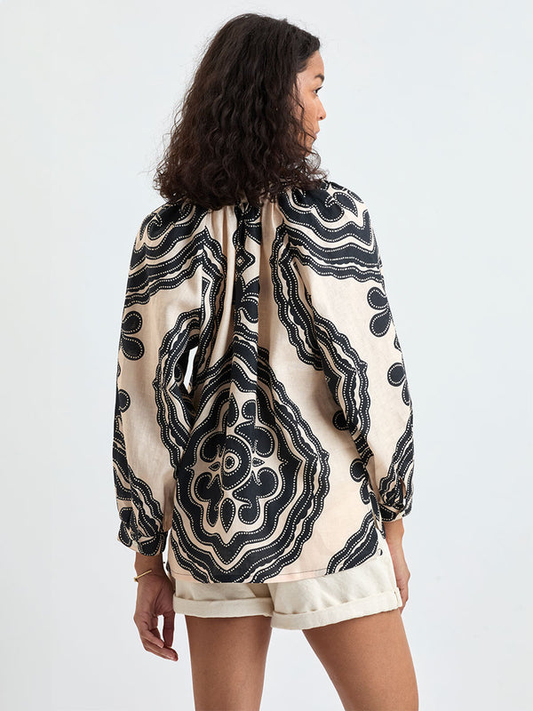 Back of a Woman Standing wearing lemlem MITA Blouse featuring ancient inspired pattern in black color on cream background and beige denim shorts