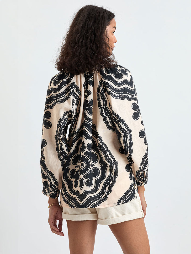 Back of a Woman Standing wearing lemlem MITA Blouse featuring ancient inspired pattern in black color on cream background and beige denim shorts