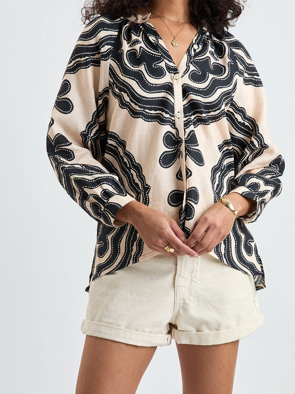 Close up on a Woman Standing wearing lemlem MITA Blouse featuring ancient inspired pattern in black color on cream background and beige denim shorts