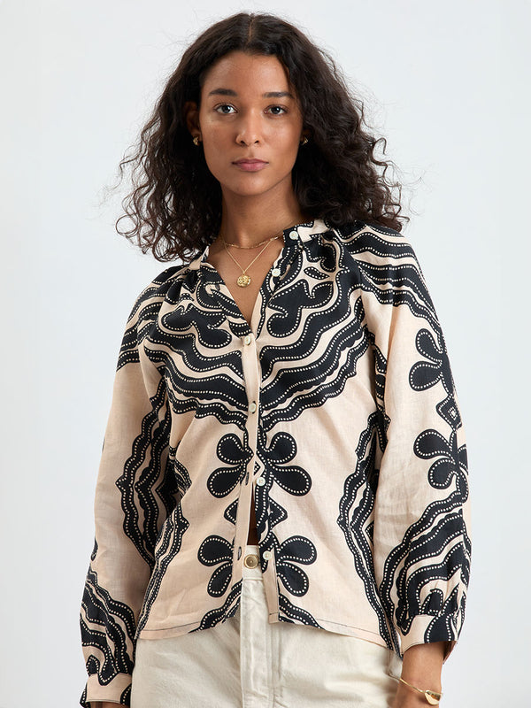 Woman Standing wearing lemlem MITA Blouse featuring ancient inspired pattern in black color on cream background and beige denim shorts