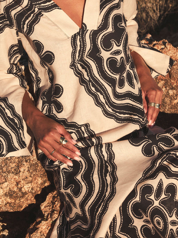 Woman sitting on stones wearing lemlem linen column dress featuring blakc pattern on cream background