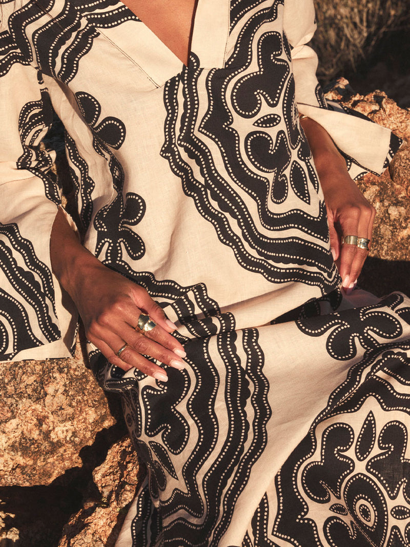 Woman sitting on stones wearing lemlem linen column dress featuring blakc pattern on cream background