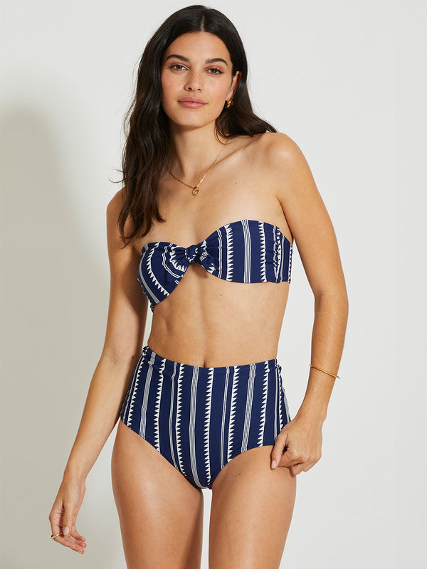 Woman Standing Wearing lemlem Bandeau Top and High Waist Bikini bottom featuring tibeb pattern in white color n navy background
