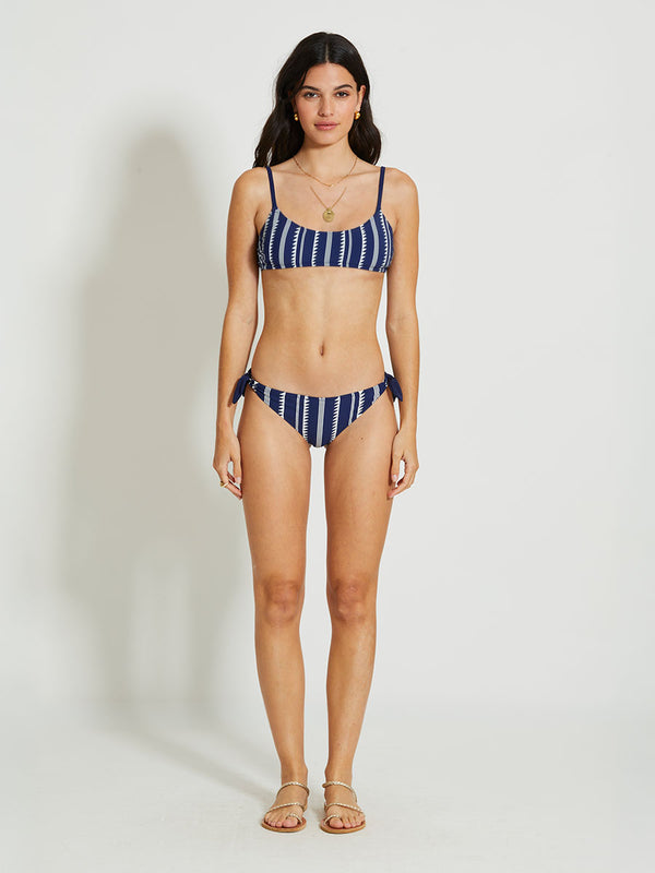 woman standing wearing a navy Nunu side tie bikini bottom with white triangles and stripes with matching bandeau top