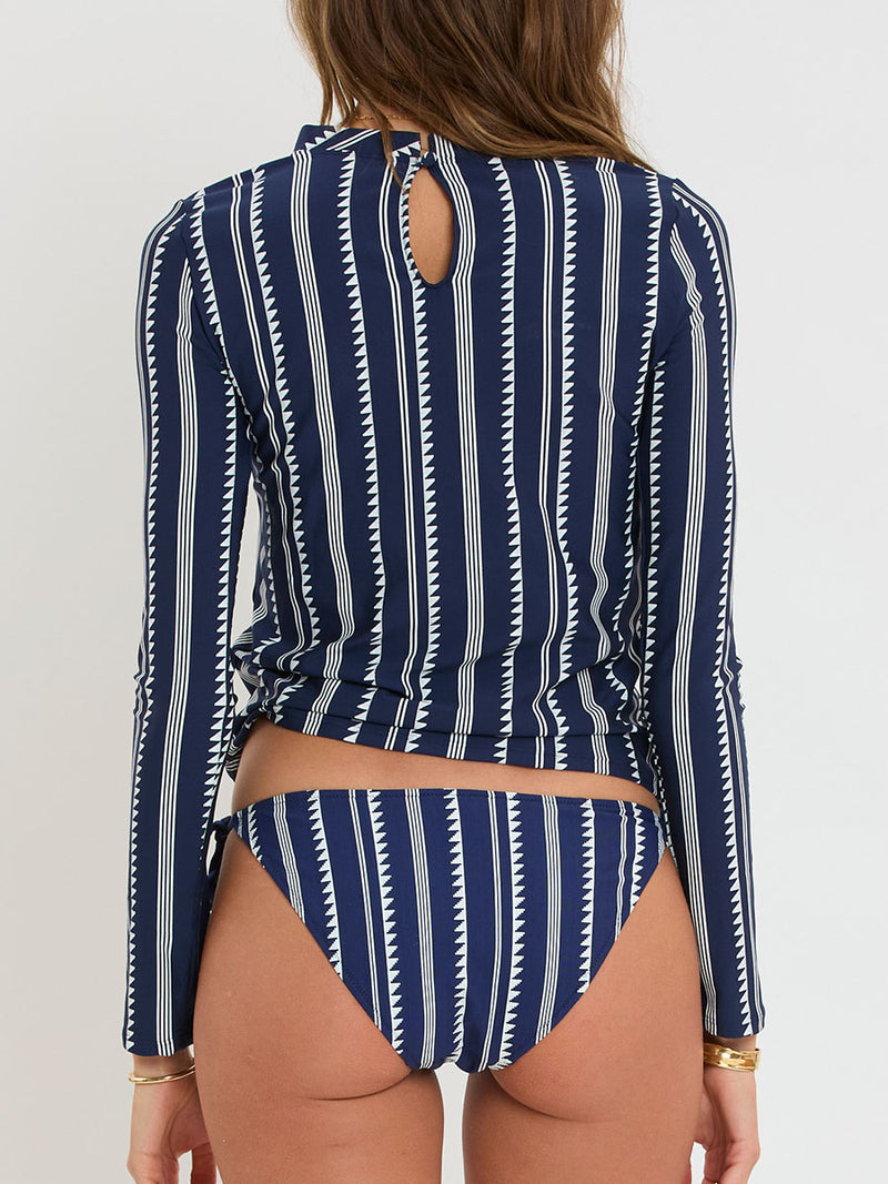 Back of a Woman standing wearing lemlem AZEB Rashguard and REKKA String Bottom featuring white tibeb pattern on navy background and handwoven MITA Blouse featuring cream color