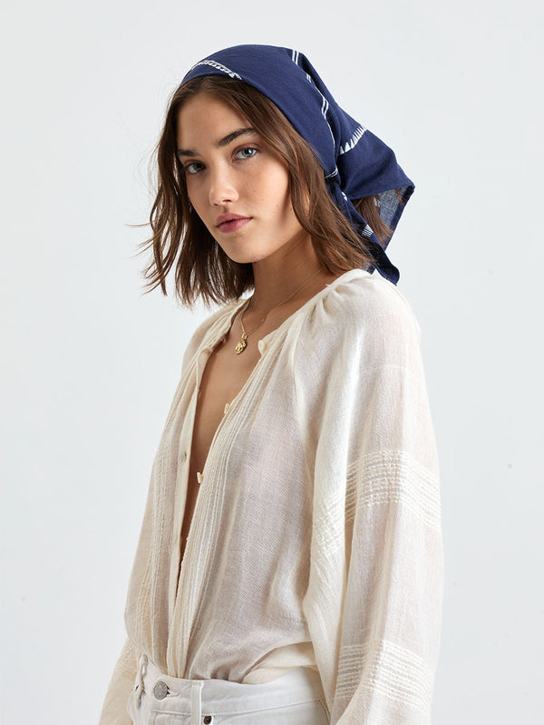 Woman standing wearing lemlem handwoven DANA Bandana featuring white tibeb pattern on navy background and handwoven MITA Blouse featuring cream color