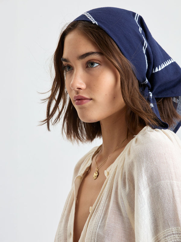 Woman standing wearing lemlem handwoven DANA Bandana featuring white tibeb pattern on navy background and handwoven MITA Blouse featuring cream color