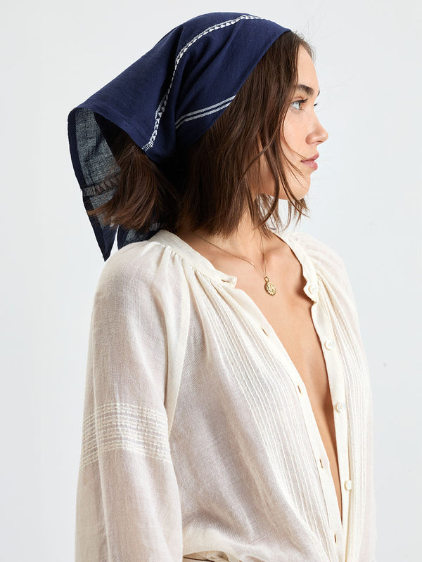 Woman standing wearing lemlem handwoven DANA Bandana featuring white tibeb pattern on navy background and handwoven MITA Blouse featuring cream color