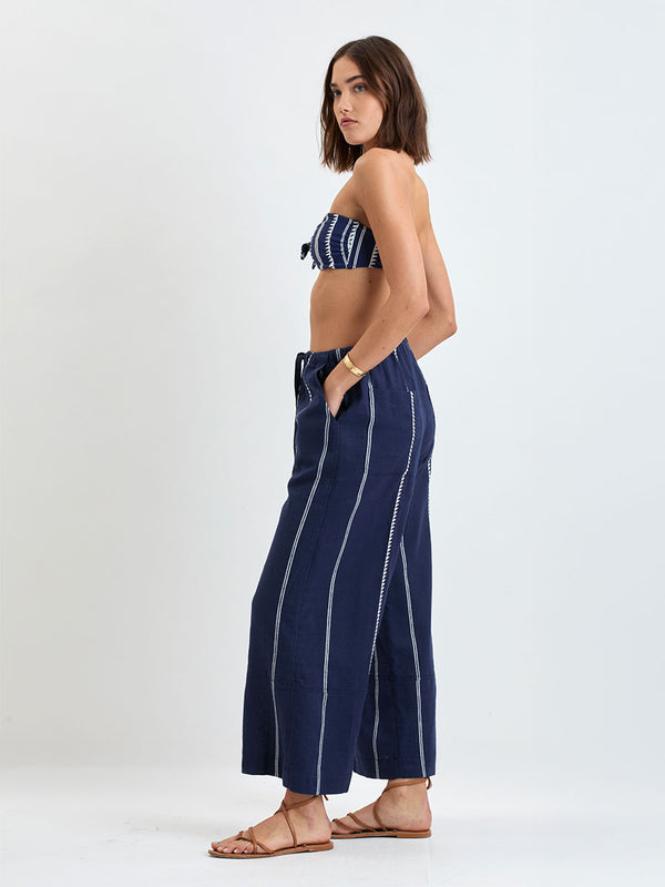 Side shot of a Woman standing wearing lemlem handwoven DESTA Pants featuring white tibeb pattern on navy background and handwoven MITA Blouse featuring cream color