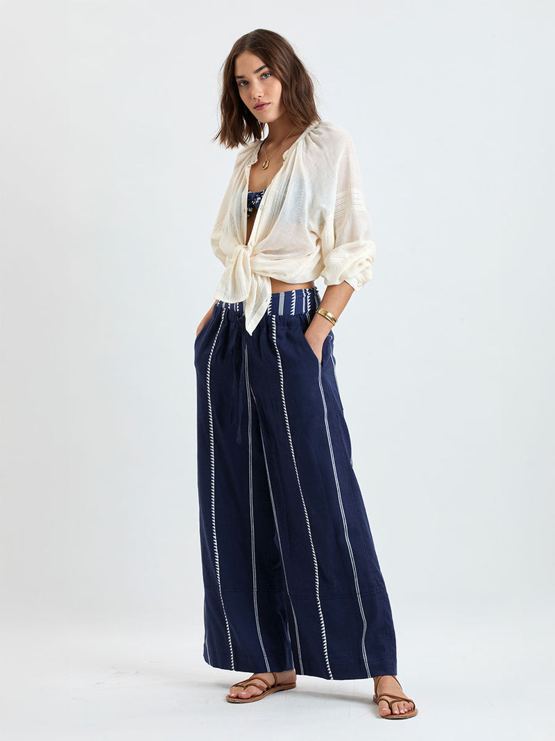 Woman standing wearing lemlem handwoven DESTA Pants featuring white tibeb pattern on navy background and handwoven MITA Blouse featuring cream color