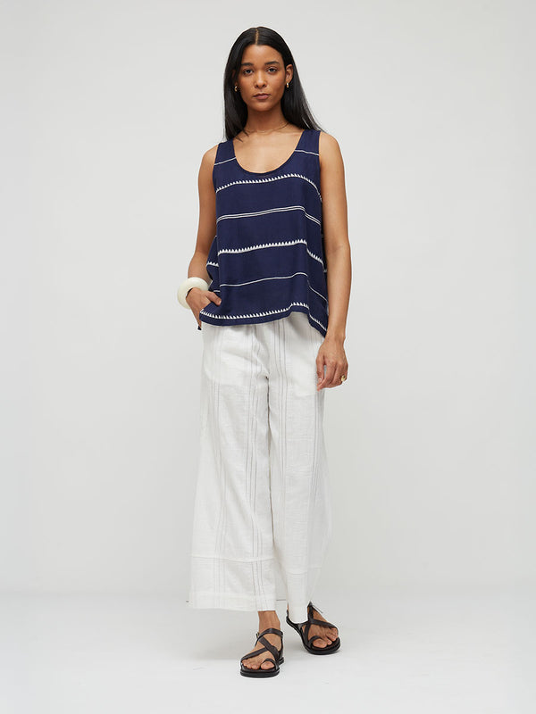 A woman modeling a lemlem handwoven KATI sleeveless navy top with thin white horizontal stripes and white wide-leg pants, standing against a light gray studio background.