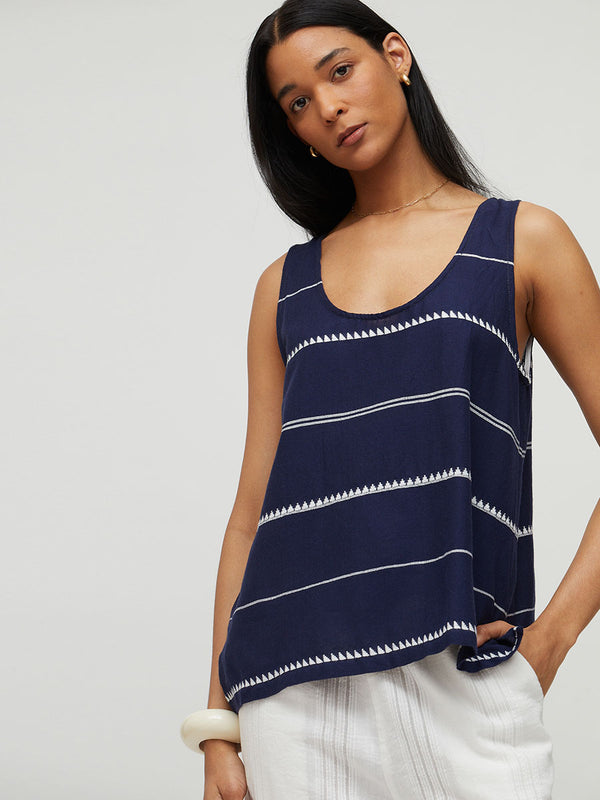 A woman wearing a lemlem handwoven KATI top with white horizontal stripes and geometric patterns, paired with white pants, against a light gray studio background.
