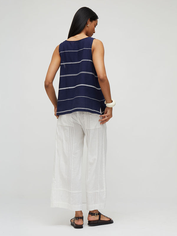 A woman shown from the back wearing a sleeveless lemlem handowven KATI  navy top with thin white horizontal stripes and white wide-leg pants, standing against a light gray studio background.