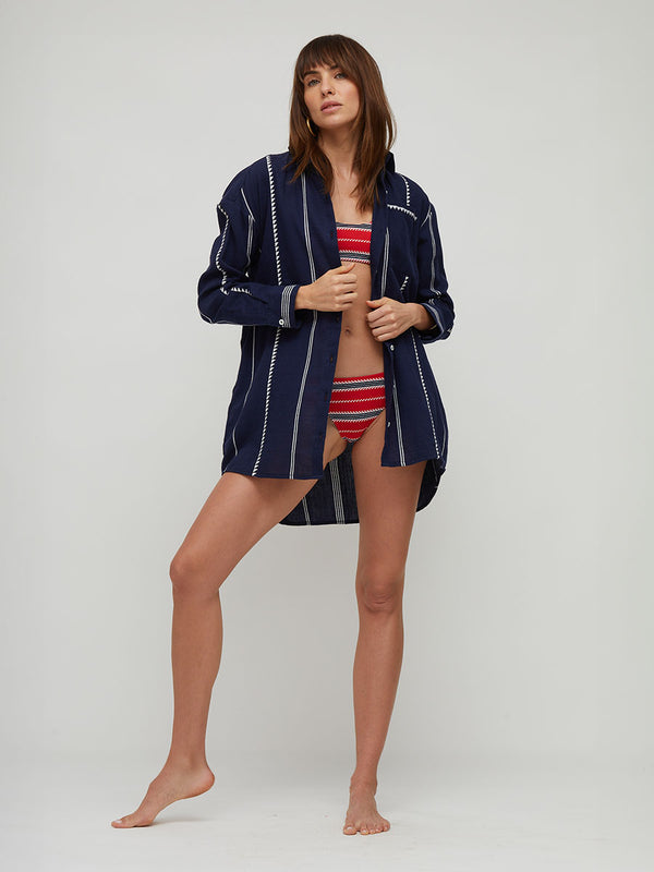 A woman modeling a navy blue open-front lemlem handwoven cover-up shirt with white vertical stripes over a red and white striped bikini, standing against a light gray studio background.