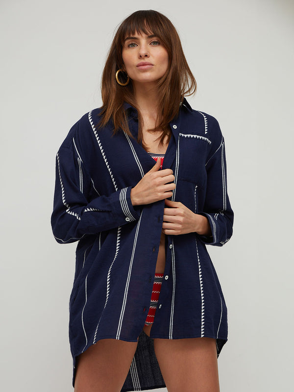A woman modeling a navy blue button-up lemlem handwoven shirt with white vertical stripes and decorative white stitched details, worn open over a red patterned top, against a plain light gray studio background.