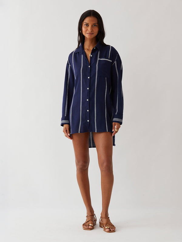 A woman modeling a navy blue button-up lemlem handwoven shirt with white vertical stripes and decorative white stitched details, worn open over a red patterned top, against a plain light gray studio background.