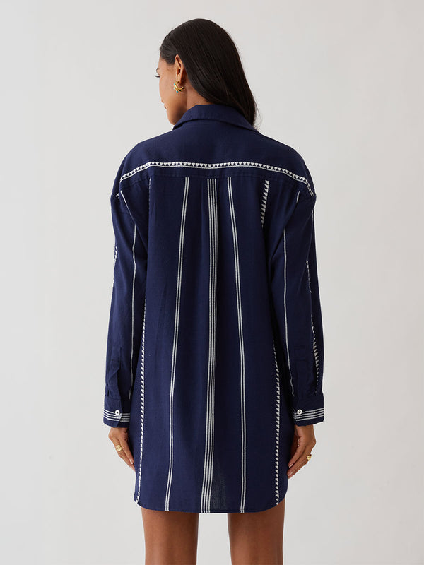 A woman modeling a navy blue button-up lemlem handwoven shirt with white vertical stripes and decorative white stitched details, worn open over a red patterned top, against a plain light gray studio background.