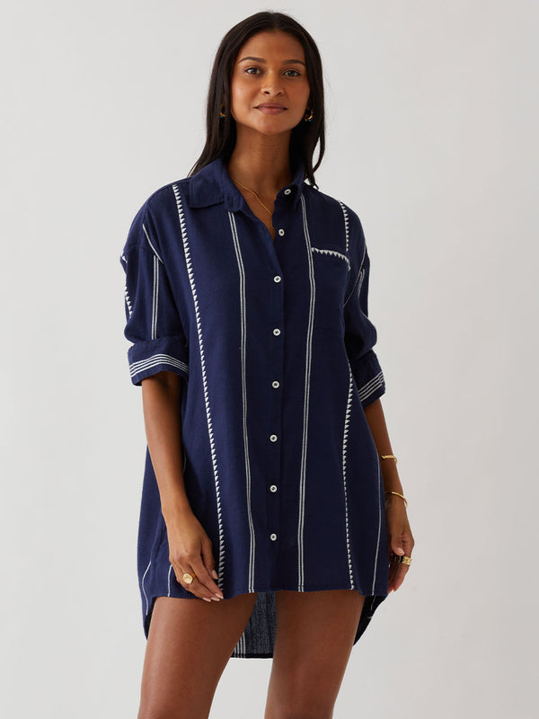 A woman modeling a navy blue button-up lemlem handwoven shirt with white vertical stripes and decorative white stitched details, worn open over a red patterned top, against a plain light gray studio background.