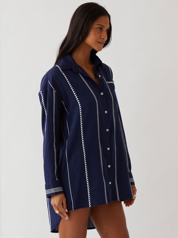 A woman modeling a navy blue button-up lemlem handwoven shirt with white vertical stripes and decorative white stitched details, worn open over a red patterned top, against a plain light gray studio background.