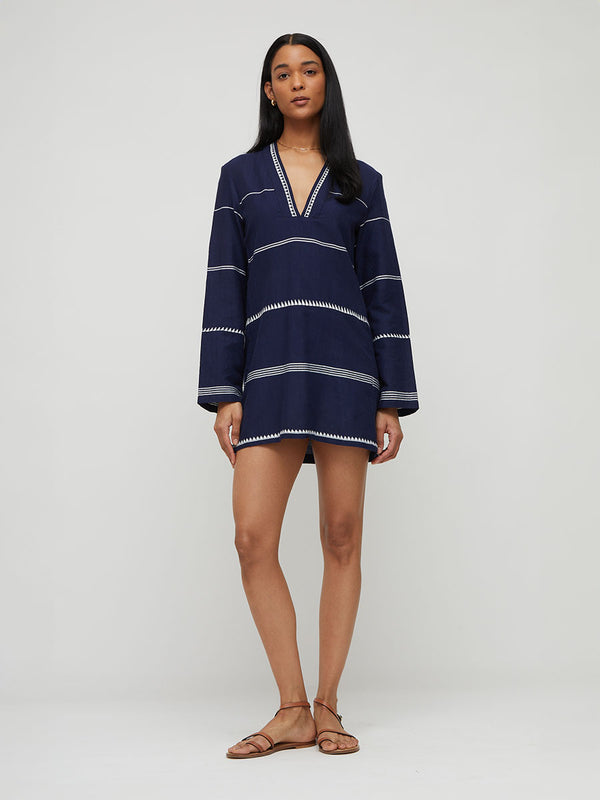 A woman modeling a navy blue long-sleeve lemlem handwoven dress with horizontal white stripes and a V-neckline, paired with brown flat sandals, against a light gray studio background.