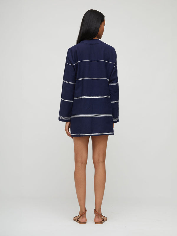 A woman viewed from the back wearing a navy blue long-sleeve lemlem hadnwoven RAEY Dress with thin white horizontal stripes and white zigzag trim along the edges, paired with brown flat sandals, against a light gray studio background.