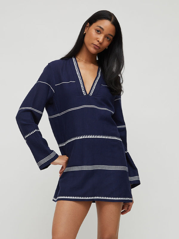 A woman modeling a navy blue long-sleeve lemlem handwoven RAEY dress with white geometric patterned trim along the V-neck, cuffs, and hem, set against a plain light gray studio background.