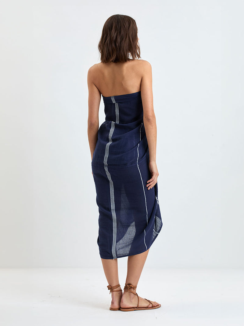 Back of a Woman standing wearing lemlem handwoven TALAKI Sarong featuring delicate white Tibeb bands on a rich navy silhouette
