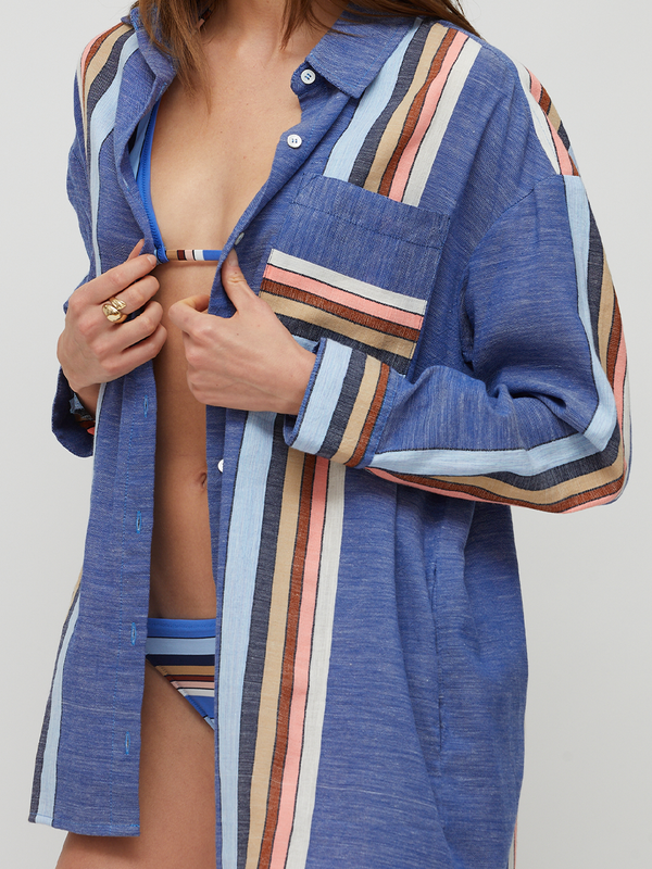 A woman wearing a loose, long-sleeve button-up lemlem handwoven MARIAM shirt in heathered blue with vertical multicolor stripes in beige, pink, brown, and white. She is opening the shirt to reveal a blue bikini underneath, set against a plain light gray studio background.