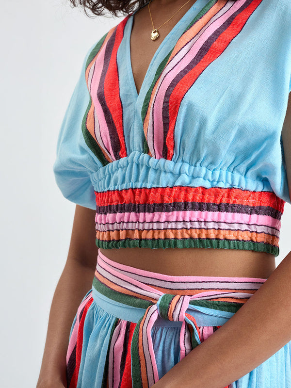 Close up on a woman wearing lemlem ALIA Plunge Top and TOLA Skirt featuring color block stripe pattern in white on sky blue background