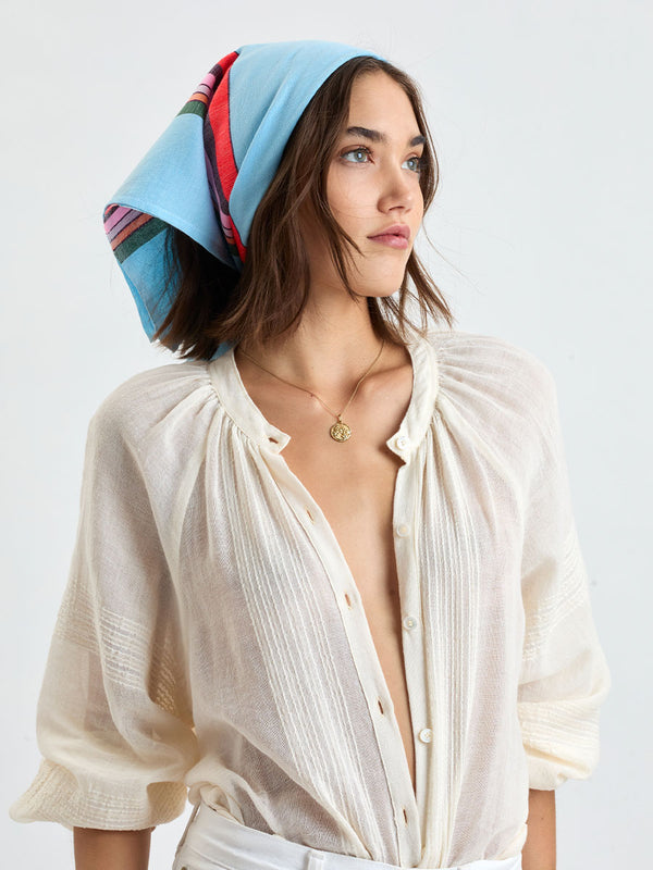 Woman wearing lemlem DANA Bandana featuring color block stripe pattern in white on sky blue background, MITA Blouse in cream color and white jeans